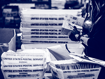 person holding stack of mailed in ballots with postal service boxes